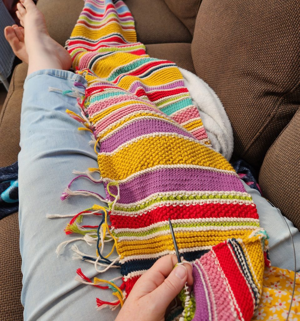 A colourful knitted scarf stretched across someone's lap, extending past their feet